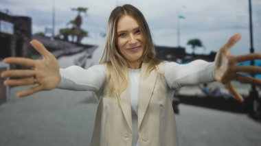 Deniz kenarındaki bir yerde gülümseyen ve kendini kucaklayan bir kadın, bulanık arka planda mavi şemsiyeler ve plaj ortamı rahatlamayı öneriyor..