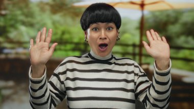 Genç Çinli bir kadın, açık hava manzarası ve neşeli bir atmosfer olduğunu düşündüren bulanık doğal bir arka plana karşı ellerini kaldırarak ve neşeli bir ifadeyle şaşkınlığını dile getiriyor..