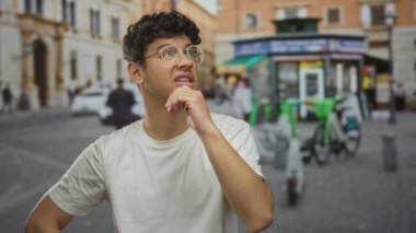 Elleri çenesinde gözlüklü bir adam caddede duruyor. Bulanık dükkanları ve park etmiş bisikletleri var. Belirsizlik düşüncesi..