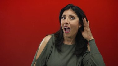 Sağ kulağını kavrayan ve stüdyoda öne doğru eğilen parlak kırmızı arka planlı bir kadın açık bir şekilde belli belirsiz bir ses duymak için; merak.