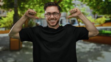 Parkta sakallı ve gözlüklü genç bir adam kendini göstererek enerjik bir şekilde çevresini çevreleyen yeşil ve ağaçlarla çevrili..