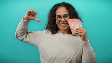 Orta yaşlı bir kadın elinde mavi arka planda Malezya bankaları tutuyor, gülümsüyor ve gururla kendisine işaret ediyor..