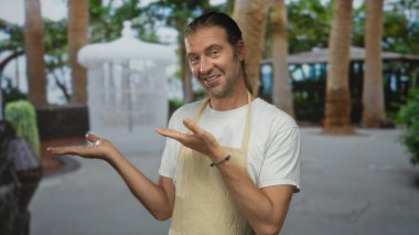 Önlüklü sarışın adam gür yeşil orman arkaplanına karşı avuçlarını açık gösterir sıcak gülümseyerek; hoş geldiniz.