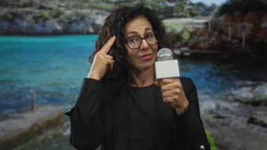 Orta yaşlı İspanyol kadın muhabir, kumsalda mikrofon tutarken parmağıyla işaret ediyor..