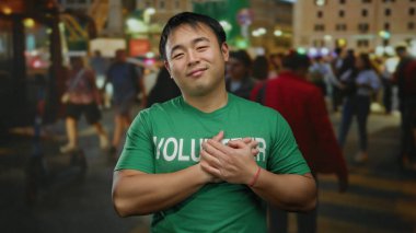 Yeşil gönüllü gömlekli genç adam, şehir sokaklarında geceleri gülümsüyor, şehir ışıkları ve bulanık yayalarla çevrili, toplum ruhunu somutlaştırıyor..