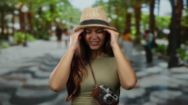 Güneşli parkta başı ağrıyan bir kadın şapkasını ve kamerasını omzuna dayayıp, bulanık yeşilliğin ortasında, yazın açık havada seyahat rahatsızlığını öneriyor.