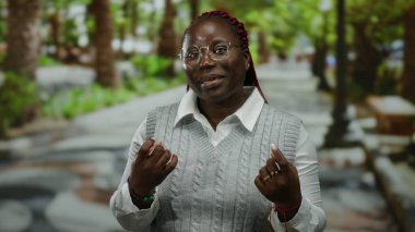 Gülümseyen ve el kol hareketi yapan bir kadın parkın içinde elleri ile davetkar ve neşeli bir hava sergiliyor.