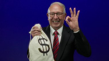 Takım elbiseli kıdemli bir adam elinde para çantası ve üzerinde dolar işareti olan mavi arka planda zenginlik ve yolsuzluğu gösteren bir işaret var..