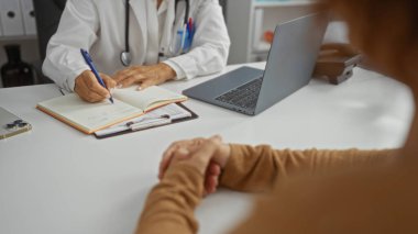 Doktor bir klinik odasında hastaya danışmanlık yaparken notlar yazıyor, sağlık sektöründeki profesyonelliği ve orta yaşlı bir İspanyol doktorla hastası arasındaki dinamiği vurguluyor..