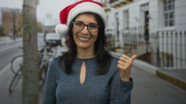 Orta yaşlı İspanyol kadın gözlüklü ve kırmızı Noel Baba şapkalı, başparmağını kaldıran ve bir şehir caddesinde gülümseyen; tatil neşesi.