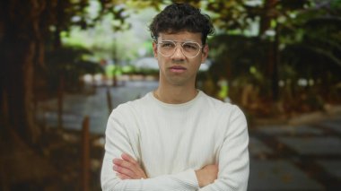 Beyaz süveter giyen ve gözlüklü bir adam ağaçlarla ve patikayla kaplı yeşil parkta kollarını kavuşturuyor..