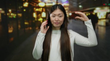 Şehir caddesinde gece vakti, arka planda bulanık ışıklarla şakacı hareketler yapan bir kadın telefonda konuşuyor..