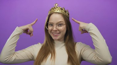 Taçlı ve gözlüklü genç bir kadın gülümsüyor ve mor stüdyoda parmağıyla tacı işaret ediyor..