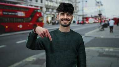 Meşgul şehir caddesinde gülümseyen genç adam sembolik kırmızı otobüsle Londra 'nın olay yerini gösteriyor, yeşil kazak giyiyor, seyahat ve şehir hayatını tasvir ediyor..