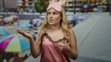 Gece elbiseli, tavşan kulaklı bir kadın renkli bir plaj arkaplanının yanında konuşuyor, gündelik ve genç yaz atmosferini vurguluyor..