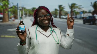 Güneşli bir caddede elinde ilaç tutarken steteskoplu gülümseyen ve puanlı Afrikalı Amerikalı kadın doktor..