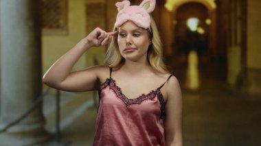 Müzede çılgın bir jest yapan kadın gece elbisesi ve uyku maskesi takıyor, sarı saç, loş ışıklandırma, oyuncu bir ifade ve eşsiz bir atmosferi olan sanat galerisi ayarları..