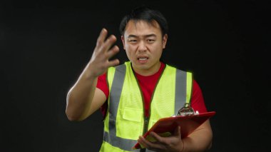 Korumalı yelek giyen genç Asyalı bir adam, siyah bir arkaplana karşı bir pano tutarken enerjik bir şekilde el kol hareketi yapar ve izole bir ortamda kararlılığını ifade eder..