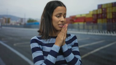 Çizgili tişörtlü genç bir kadın arka plandaki nakliye konteynerleriyle ilgili endişelerini dile getiriyor..