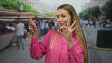 Pembe tişörtlü bir kadın şehir pazarında gülümseyip işaret ederken insanların ve tezgahların bulanık arkaplanlarını gösteriyor..