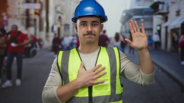 Güvenli yelek ve şapka giyen genç İspanyol adam, kalabalık şehir caddesinde yeminli bir şekilde el kaldırıyor. Şehir hayatının ortasında sorumluluk ve kararlılığı yansıtıyor..