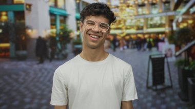 Kıvırcık saçlı ve gözlüklü bir adam başını yana çevirir ve kalabalık bir şehir caddesinde gülümser, beyaz bir tişört giyer; mutluluk.