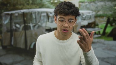 Gözlüklü genç adam, kampçı tarafından desteklenen yeşil parktaki caddede kamerayı işaret ediyor..