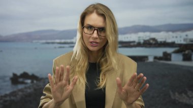 Elleri deniz kenarında sakince el sallayan, rüzgarda hafifçe savrulan sarı saçlı, gözlüklü ve kahverengi ceketli, gün ışığında sakin bir okyanus zeminine yaslanan bir kadın..