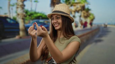 Açık havada gülümseyen bir kadın, güneşli bir sokakta, palmiye ağaçları içinde, şapka takıyor ve tatil sırasında neşeli turist tavırları sergiliyor..