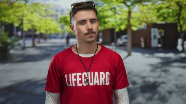 Genç İspanyol cankurtaran şehir caddesinde görünür bir şekilde dalgın, cankurtaran yazılı kırmızı bir tişört giymiş, arka planda ağaçlar olan parlak güneş ışığı altında duruyor..