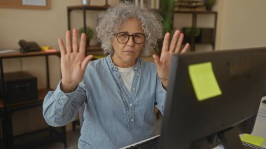 Ofis ortamında el kol hareketi yapan, orta yaşlı, kıvırcık saçlı, gözlüklü ve kot gömlekli, bilgisayarın önünde el kaldırarak iletişim hareketi yapan bir kadın..