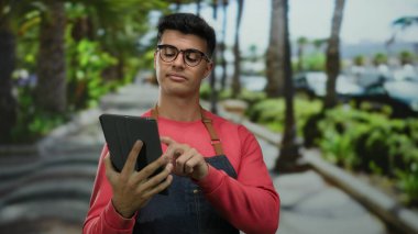 Gözlüklü genç adam dışarıda, yemyeşil bir park ortamında tablet kullanıyor, günlük kırmızı kıyafetler ve önlük giyiyor, odaklanmış ve dokunmatik ekran cihazıyla meşgul görünüyor..