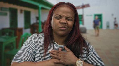 Yeşil binanın girişinde göğsünü sıkıca tutan kadın çizgili gömlek ve tahta kol saati takıyor; kalp krizi acısı.