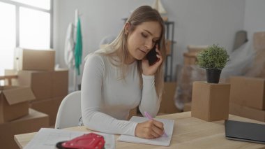 Yeni bir evde notlar alırken telefonla konuşan bir kadın oturma odasındaki kutuları taşıyarak pencerelerden içeri doğal ışık giriyor..