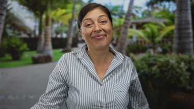 Güneşli bir günde palmiye ağaçlarının altındaki yemyeşil parkta gülümseyen orta yaşlı İspanyol kadın çizgili gömlek giyiyor..