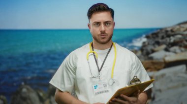 Deniz kenarındaki panoya bakan sakallı genç doktor. Arka planda okyanus var. Açık havada steteskop takıyor.