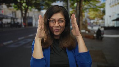 Mavi ceketli kadın, elleri ağaçlarla dolu bir caddede yüzünü çerçeveliyor..