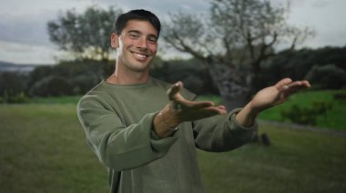 Yeşil süveter giyen genç adam, parkta gülümseyerek göğsüne bastırıyor..