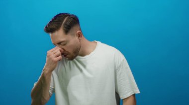 Sakallı genç adam yorgun görünüyor, burnu mavi bir arka plana sürtünüyor, izole edilmiş, günlük beyaz gömlek giyiyor, yorgunluk veya stres taşıyor..