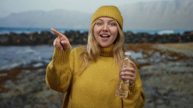 Sarı kazaklı ve şapkalı kadın deniz kenarına neşeli bir ifadeyle bakarken şişeyi tutuyor, plaj manzarasına karşı duruyor, rahat bir açık hava anı yakalıyor..