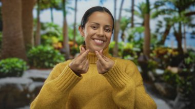 Hardal rengi kazaklı kadın parmaklarıyla kalp hareketi yapıyor yemyeşil bir parkta palmiye ağaçları ve açık hava ortamını güzelleştiren canlı bitkiler.
