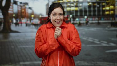 Parlak turuncu hapishane tulumlu kadın kalabalık şehir caddesinde duruyor, akşam üstü aydınlığında bulanık şehir geçmişiyle dışarıda duygularını ifade ediyor..