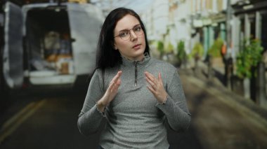 Arka planda teslimat minibüsüyle kalabalık bir caddede duran, gözlüklü, gri kazaklı, esmer saçlı, kentsel çevreyle uyumlu, hareketli şehir yaşamını öneren bir kadın..