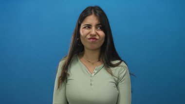 İzole edilmiş mavi arka plan duvarına karşı duran genç İspanyol kadın, bir portre ortamında düşünceli bir ifadeyle, tarafsız bir çevrede yansıtıcı bir mizacı somutlaştırıyor..