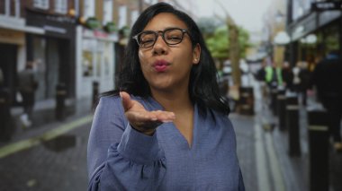 Mavi bluz giyen ve gözlüklü bir kadın gün ışığında dudaklarıyla öpücük yolluyor..