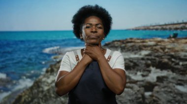 Kayalık sahil kenarında gözleri kapalı, elleri boynuna dokunmuş, önlük giymiş, arka planda okyanus ve gökyüzü olan bir kadın, sakin bir tefekkür çağrıştırıyor..