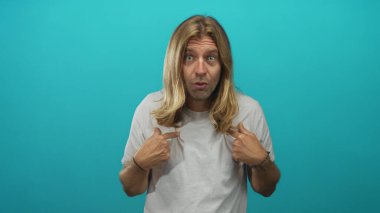 Sarışın, uzun saçlı, turkuaz bir stüdyo stüdyosunda parmaklarını göğsüne gösteren, şaşırmış bir yüz ifadesi olan, sürpriz kişilik keşfi yapan bir adam..