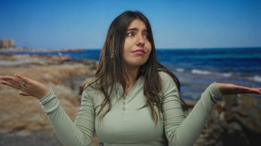 Günlük giysili bir kadın, deniz kenarında, kayalık sahiller ve okyanus zeminiyle kafası karışmış, yeşil bluz giymiş ve güneşli bir günde tropikal plajda avuçlarını açık tutarak el sallıyor.