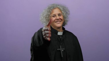 Papaz kıyafeti giymiş bir kadın, mor arka plana karşı elini uzatıyor. Gri saçlı, siyah giysili ve haçlı kolyeli, dindar ve misafirperver biri..