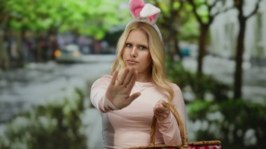 Açık havada tavşan kulağı takan genç sarışın bir kadın elinde sepet tutarken elinde bir şehir sokağında komik jestler yapıyor.